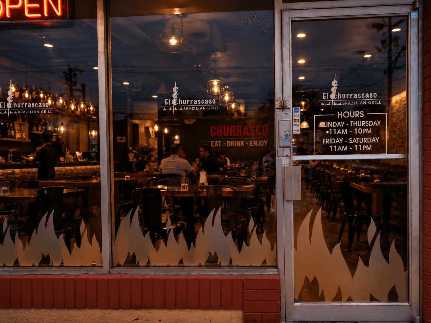 El Churrascaso storefront glass at dusk with flame-shaped frosted vinyl graphics, an OPEN neon sign and logo and hours decals on the door