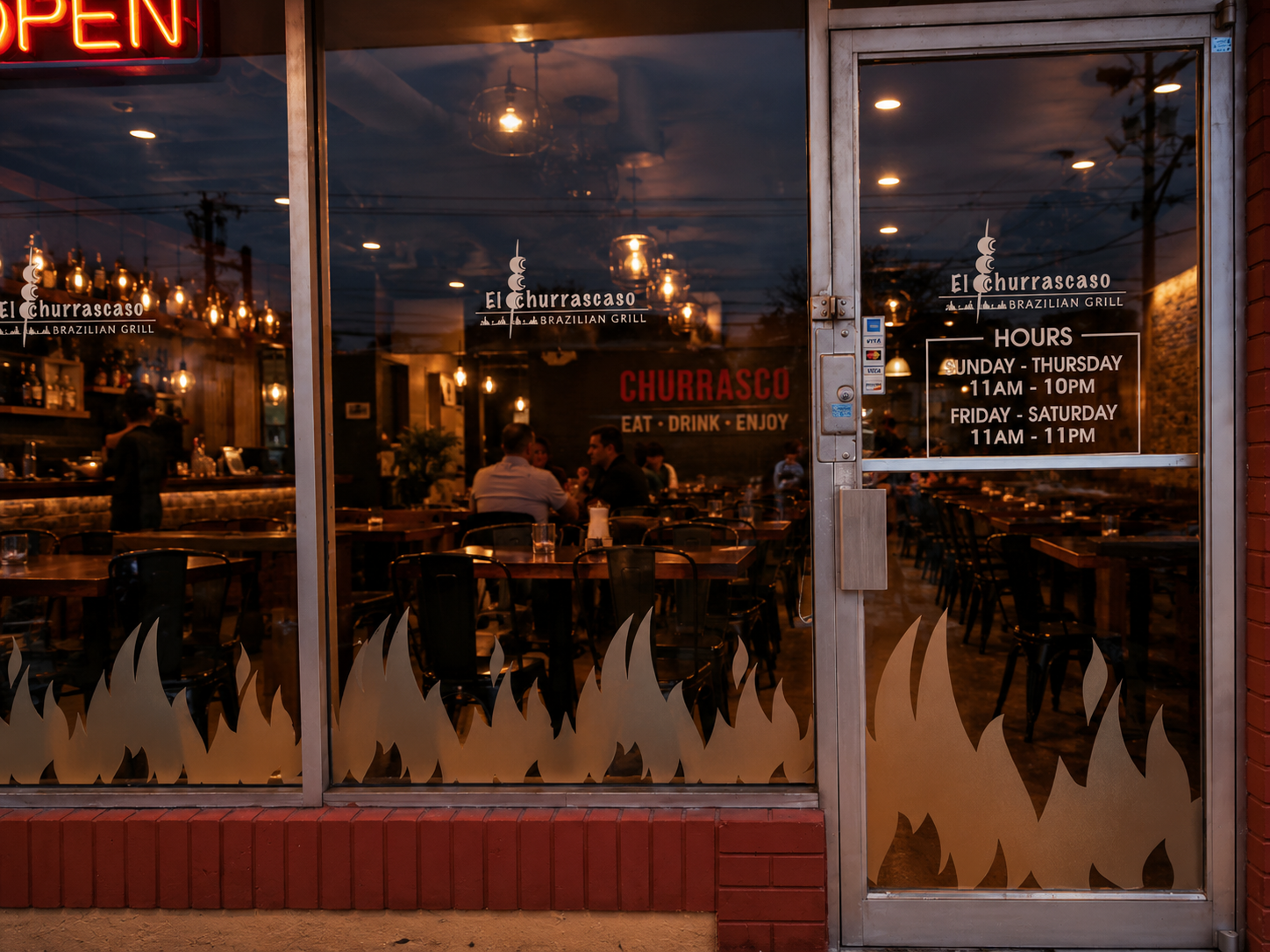 El Churrascaso storefront glass at dusk with flame-shaped frosted vinyl graphics, an OPEN neon sign and logo and hours decals on the door
